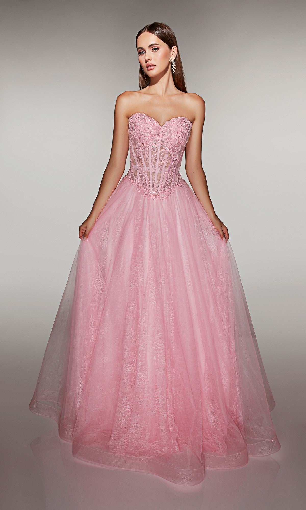 A woman in the Alyce 61984 long prom dress, a light pink strapless tulle ball gown with a corset bodice and lace details, stands against a neutral background.